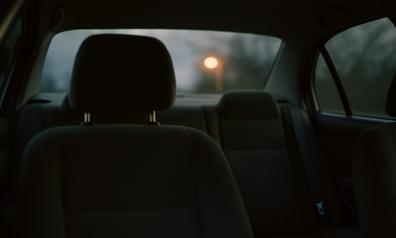 The inside of a parked sedan at dusk seen from the back seat, the driver's headrest in shadow and a single orange streetlight glowing through the rear window